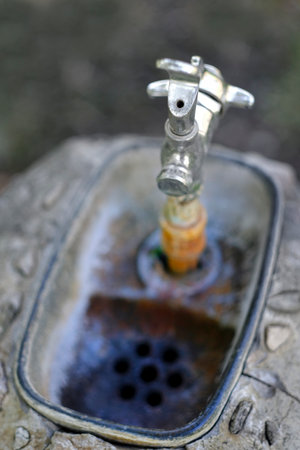 Old worn drinking fountain with handle in park with green grass and drainの写真素材