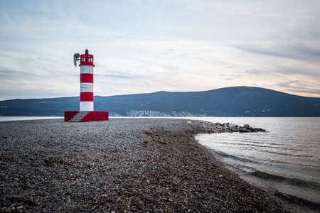 curved pebble beach with red and white striped lightouse with hills in the background in Kotor bayの写真素材