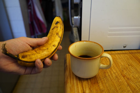 A man is holding a banana and a cup of coffee in the kitchen.の写真素材