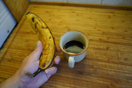 Man's hand holding a cup of coffee and a banana on a wooden tableの写真素材