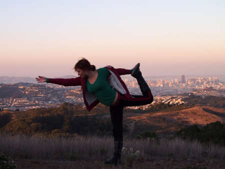 Beautiful Lady Does Lord of the Dance Pose I - Natarajasana with San Francisco cityscape in the distance at dusk.の写真素材