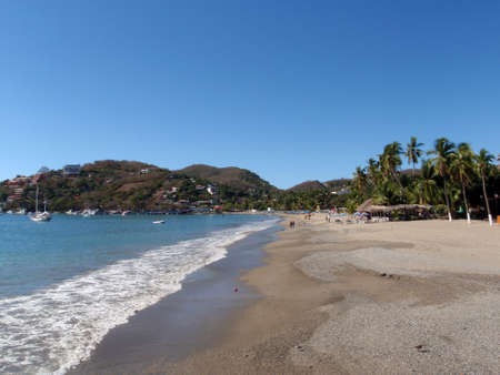 Beautiful tropical sandy beach with boats in the water and hills in the background in Zihautanejo, Mexicoの写真素材