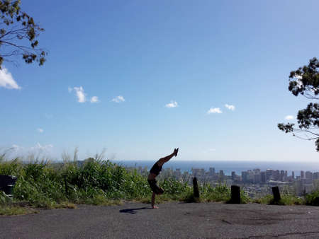 Man wearing t-shirt, shorts and rubber slippers Handstanding with a curved back at Tantalus lookout point with the city of Honolulu, Hawaii and ocean visible in the distance.の写真素材