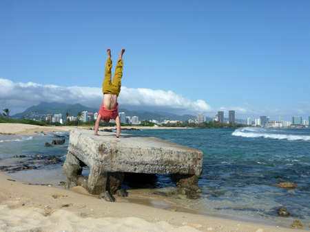 Man wearing a t-shirt, pants, and watch Handstands on old pillbox on Sand Island beach with the City of Honolulu in the distance.の写真素材