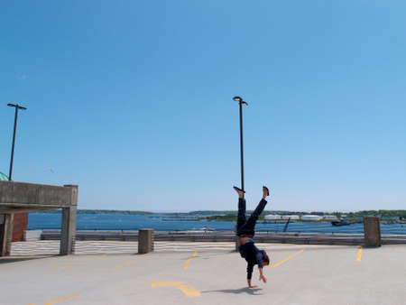 Man wearing a hoodie, pants, and slippers does one handed Handstands on top of a parking garage the City of Portland Maine with Casco bay in the distance.の写真素材