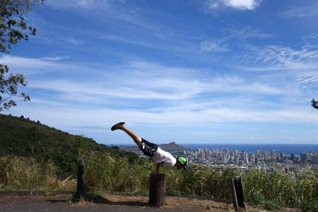Man wearing hat, t-shirt, shorts, and slippers does balances on hands as he does Mayurasana or...