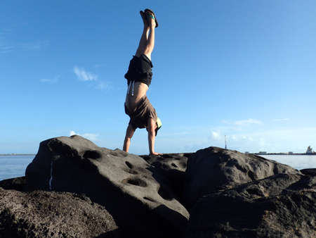 Man Handstands on rock jetty in Kahului Harbor wearing a hat, t-shirt, shorts, and slippers with his...