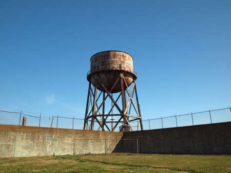 Rusting water tower stands beyond the wall  and bard wire fence of grassy Alcatraz's prison yard.のeditorial素材