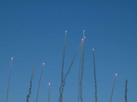 Red Fireworks soar into the air trailed by smoke during the day against a blue skyの写真素材