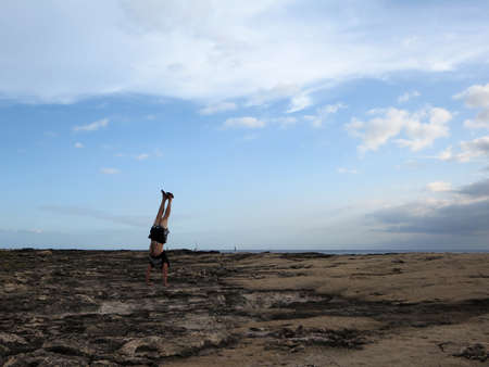 Man wearing a t-shirt, shorts, and slippers Handstands on shore rocks of Ko Olina on Oahu, Hawaii with dramatic clouds, sky, and pacific ocean in the distance.の写真素材