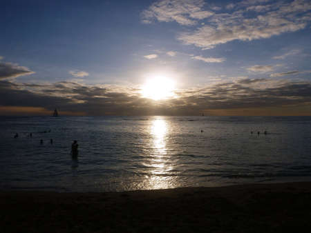 People hang in the water as a dramatic sunset happens over the clouds and reflecting on the Pacific ocean on the water of Waikiki as boat sails in water on Oahu, Hawaii.の写真素材