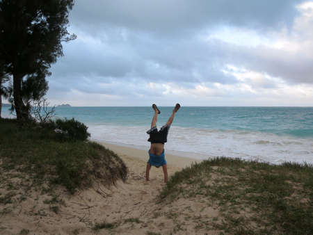 Man does handstand in the sand wearing t-shirt and shorts on the Waimanalo beach on Oahu with his legs open with a cloudy sky.の写真素材