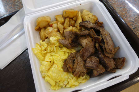 Kiawe grilled steak, scrambled eggs, country fried potatoes in a styrofoam plate on a tray on a table.の写真素材