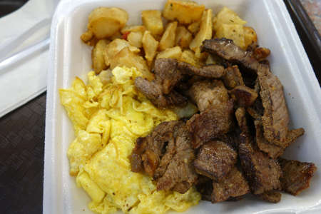 Kiawe grilled steak, scrambled eggs, country fried potatoes in a styrofoam plate on a tray on a table.の写真素材