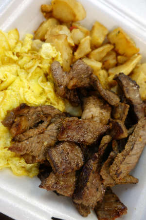 Kiawe grilled steak, scrambled eggs, country fried potatoes in a styrofoam plate on a tray on a table.の写真素材