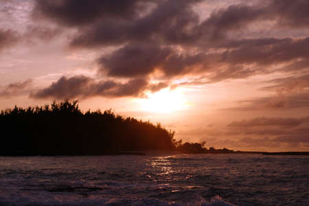 Sunset through the clouds and over the trees as it reflects on the watery waves at Kawela Bay on the North Shore of Oahu.の写真素材