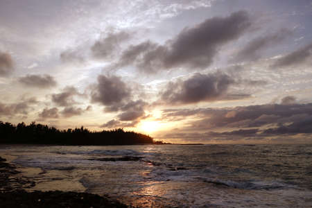 Sunset through the clouds and over the trees as it reflects on the watery waves at Kawela Bay rocky shore on the North Shore of Oahu.の写真素材
