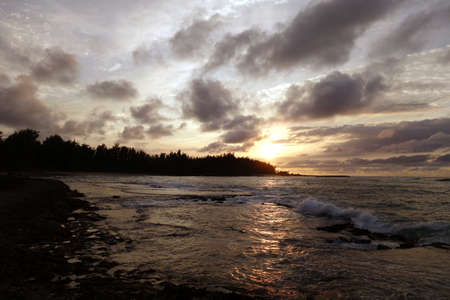 Sunset through the clouds and over the trees as it reflects on the watery waves at Kawela Bay rocky shore on the North Shore of Oahu.の写真素材
