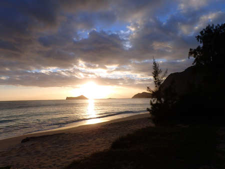 Early Morning Sunrise on Waimanalo Beach on Oahu, Hawaii in between Rabbit and Rock Island bursting through the clouds and trees on the shore.  February 2015.の写真素材