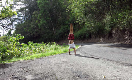 Man wearing a hat, t-shirt, shorts, and slippers Handstands at Tantalus Mountain road with lush vegetation above the City of Honolulu on Oahu, Hawaiiの写真素材