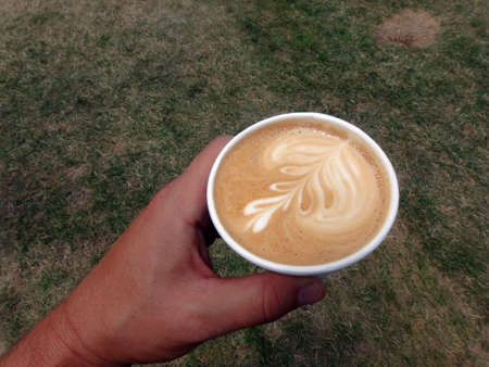 Hand holding a styrofoam cup of Cappuccino with a leaf pattern on grass field.の写真素材