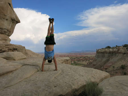Man wearing a hat, t-shirt, shorts, and slippers does Handstand on cliffs with field landscape in the desert Mountain with dramatic clouds in sky in Utah, USA.の写真素材