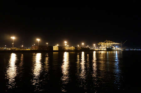 OAKLAND - OCTOBER 12: Fully loaded Shipping Cargo Boat unloaded at night under giants unloading cranes in Oakland Harbor with lights reflecting on the water and matson shipping containers on the shore.  Oakland California on October 12, 2015.のeditorial素材