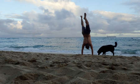 Man Handstanding on Diamond Head beach with black dog next to him as wave roll on to shore and boat...