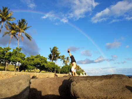 Man Handstanding on coastal rocks with Rainbow overhead and coconut trees along path on Oahu, Hawaii.                    の写真素材