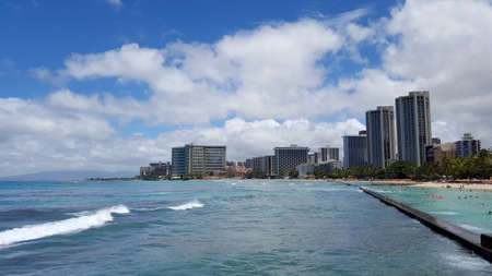 Waikiki - July 3, 2016: Waves roll into shore as People play in the protected water and hang out on the beach in world famous tourist area Waikiki on a beautiful day with hotels in the distance.  June 17 2014 in Waikiki, Hawaii.の写真素材