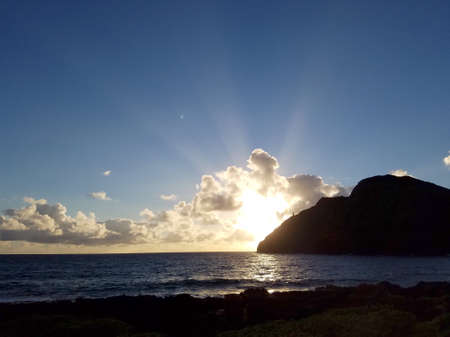 Ocean waves lap on Makapuu beach as sunrises through the clouds with Makapuu Lighthouse and point in the distance looking out to sea on the east side clouds in sky of Oahu, Hawaii.の写真素材