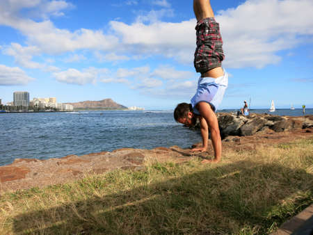 Man wearing shorts, shirt, and shoes Handstanding along shore of Magic Island with Ala Wai Boat Harbor, Waikiki, Diamond Head in the distance looking into the pacific ocean on Oahu, Hawaii on a beautiful day.の写真素材