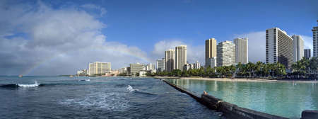 Waikiki - January 8, 2020: People play in the protected water and hang out on the beach with rainbow in world famous tourist area Waikiki on a beautiful day with hotels in the distance.のeditorial素材