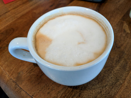 White cup of Cappuccino with a Large white dot of foam on wood table.の写真素材
