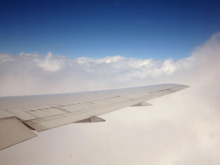 An aerial view of fluffy white clouds as seen from the window of a commercial jet plane, flying high in the sky. The wing of the plane can be seen in the image.の写真素材