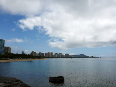 Stunning view of Honoluluâs skyline and the iconic Diamond Head crater from Ala Moana Beach Park, a popular recreational area in Hawaii. The photo was taken from the rock wall near Kewalo Basin, a harbor for fishing and recreational boats. The ocean is clear and blue, and the sky is partly cloudy.の写真素材
