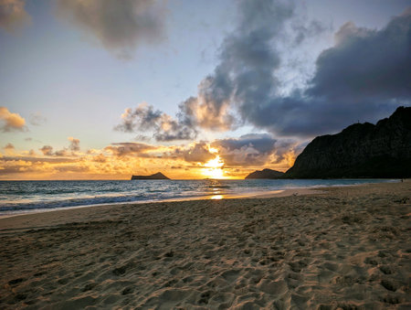 Sunrise on Waimanalo Beach, a long and sandy beach on the eastern coast of OÊ»ahu, Hawaii. The photo was taken from the perspective of someone standing on the beach, looking at the orange sun rising over the calm ocean. The sky is a mix of orange, pink, and blue colors, and there are a few clouds in the sky.の写真素材