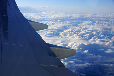 View from an airplane window, focusing on the aircraftâs wing that is visible against a backdrop of clouds and sky. The photo shows the grey wing with smooth surfaces and streamlined design, and the vast expanse of fluffy white clouds scattered across the blue sky.の写真素材