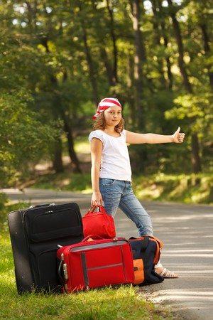 Young hitch-hiker girl standing on road side afternoon in forest with bagsの写真素材