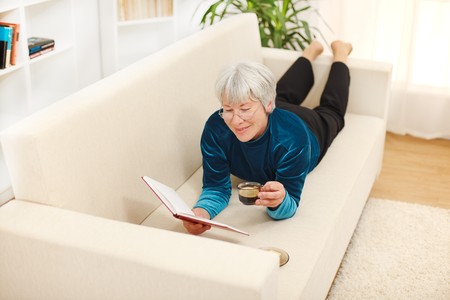 Senior woman laying on sofa, drinking coffe and reading bookの写真素材