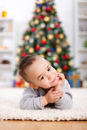 Cute boy laying in front of decorated christmas tree, prop his headの写真素材