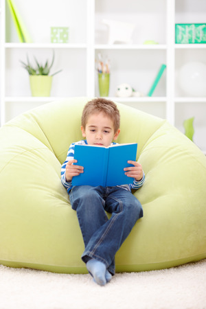 Little elementary schoolboy sitting in chair and reading from bookの写真素材