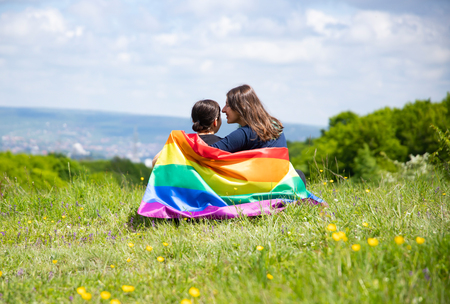 Lesbian couple sitting in a green meadow holding a Gay Rainbow Flag. Bisexual,gay, lesbian, transsexual symbol. Happiness, freedom and love concept for same sex couplesの写真素材