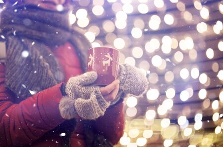 Young woman in winter gloves holding a mug of hot tea or wine in snowfall. Christmas lights in the backgroundの写真素材