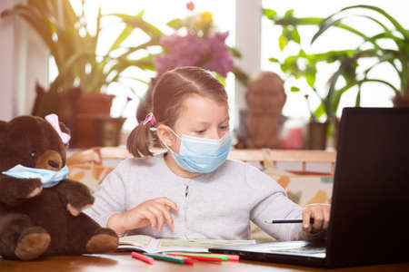 Distance learning-online education. School girl with face mask watching online education classes and doing school homework. COVID-19 pandemic forces children online learning.の写真素材