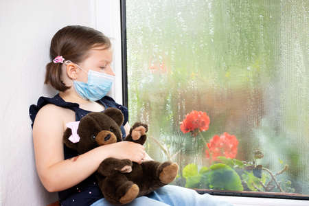 Stay at home-quarantine coronavirus pandemic prevention. Sad girl and her bunny both in protective medical masks sits on windowsill and looks out window. Prevention epidemic concept.の写真素材