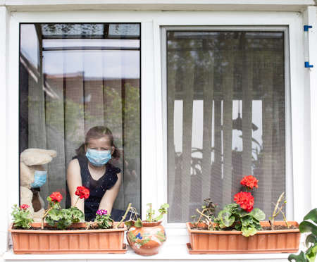 Stay at home-quarantine coronavirus pandemic prevention. Sad girl and her teddy bear both in protective medical masks sits on windowsill and looks out window. Prevention epidemic concept.の写真素材