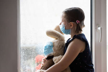 Stay at home-quarantine coronavirus pandemic prevention. Sad girl and her bunny both in protective medical masks sits on windowsill and looks out window. Prevention epidemic concept.の写真素材