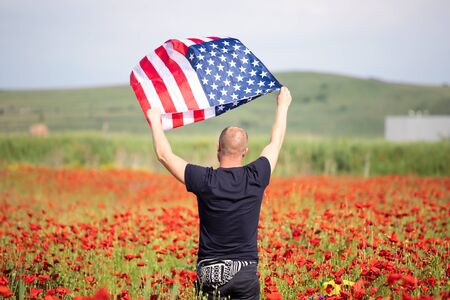 Attractive man holding Flag of the United States in beautiful poppy field on a clear, sunny day. Celebrating Independence Day, National holiday concept.の写真素材