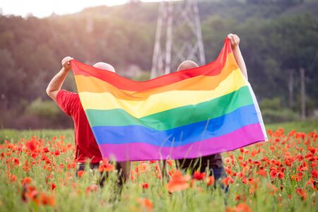 Gay couple holding a Gay Rainbow Flag over beautiful poppy field. Bisexual,gay, lesbian, transsexual symbol. Happiness, freedom and love concept for same sex couplesの写真素材